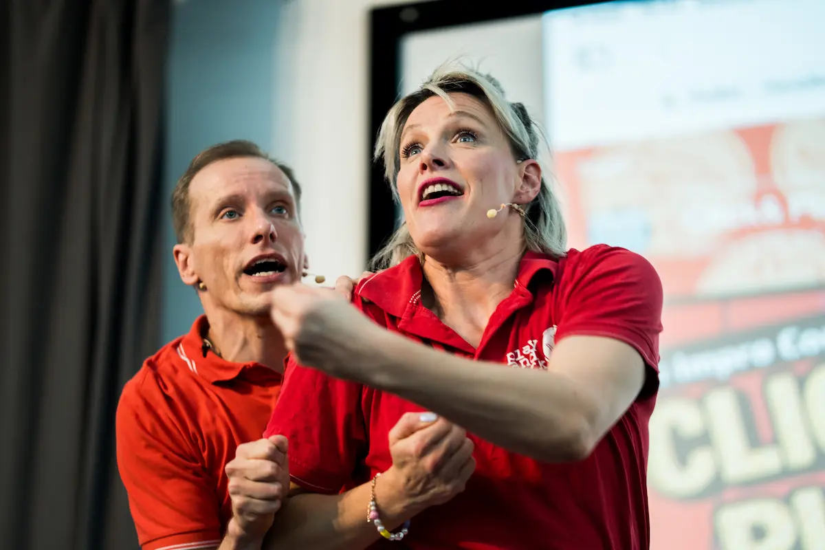 Weibliche Performerin in rotem Shirt zeigt begeistert nach vorne, männlicher Kollege dahinter mit gespannter Mimik bei der KI-Impro-Show