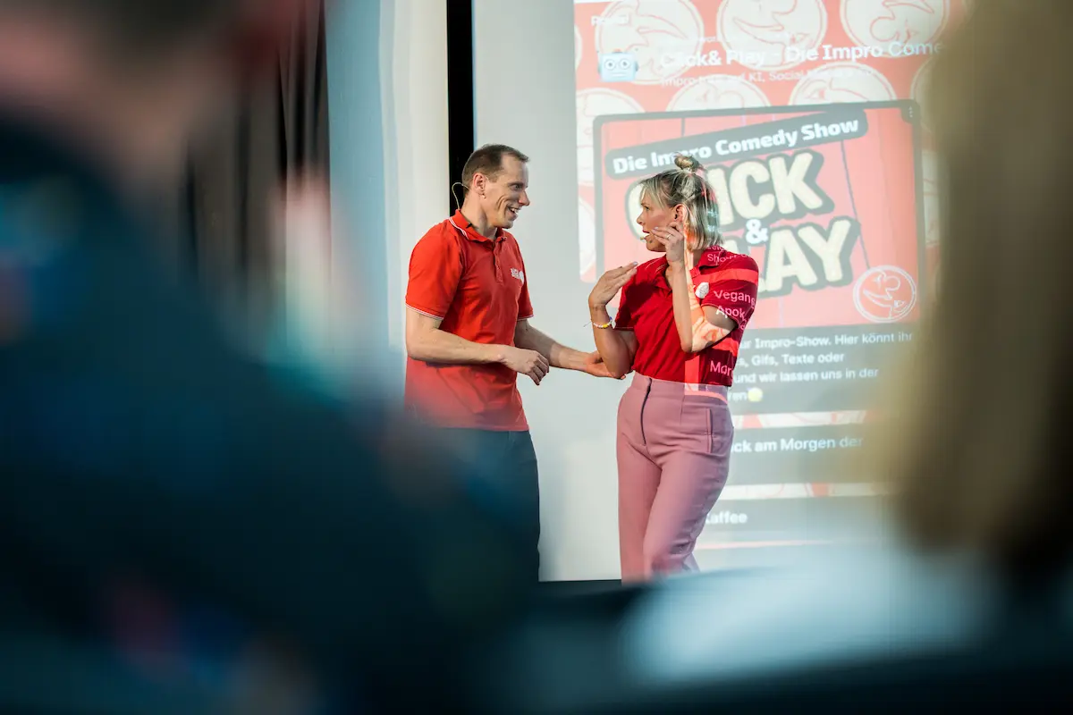 Zwei Impro-Performer in roten playandwork-Shirts bei einer humorvollen, interaktiven Szene auf der Bühne der Click & Play Impro Comedy Show in Berlin